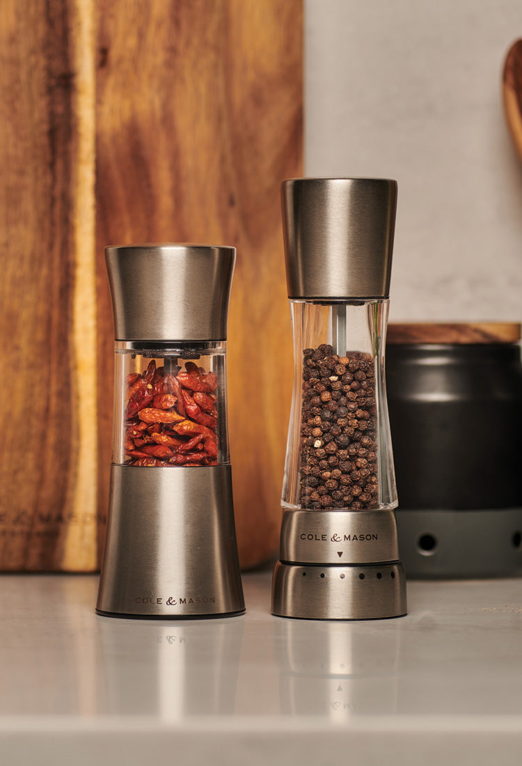Two stainless steel and glass spice grinders sit on a kitchen counter; the left grinder contains dried red chili peppers, and the right grinder contains whole black peppercorns. A wooden cutting board stands in the background.