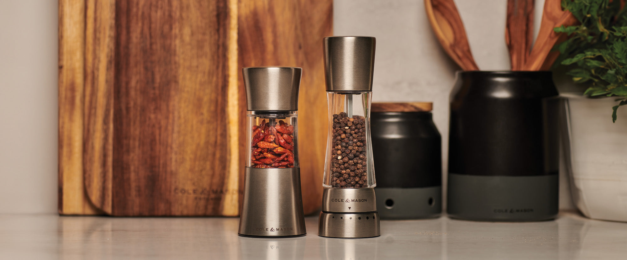 Two stainless steel spice grinders filled with dried chili peppers and black peppercorns are placed on a kitchen counter, with wooden cutting boards and kitchen utensils in the background.