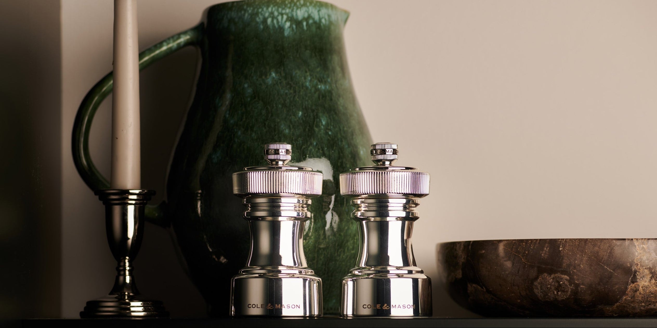 A green ceramic pitcher, a white candle in a holder, two metallic salt and pepper grinders, and a wooden bowl are arranged on a dark shelf against a beige wall.
