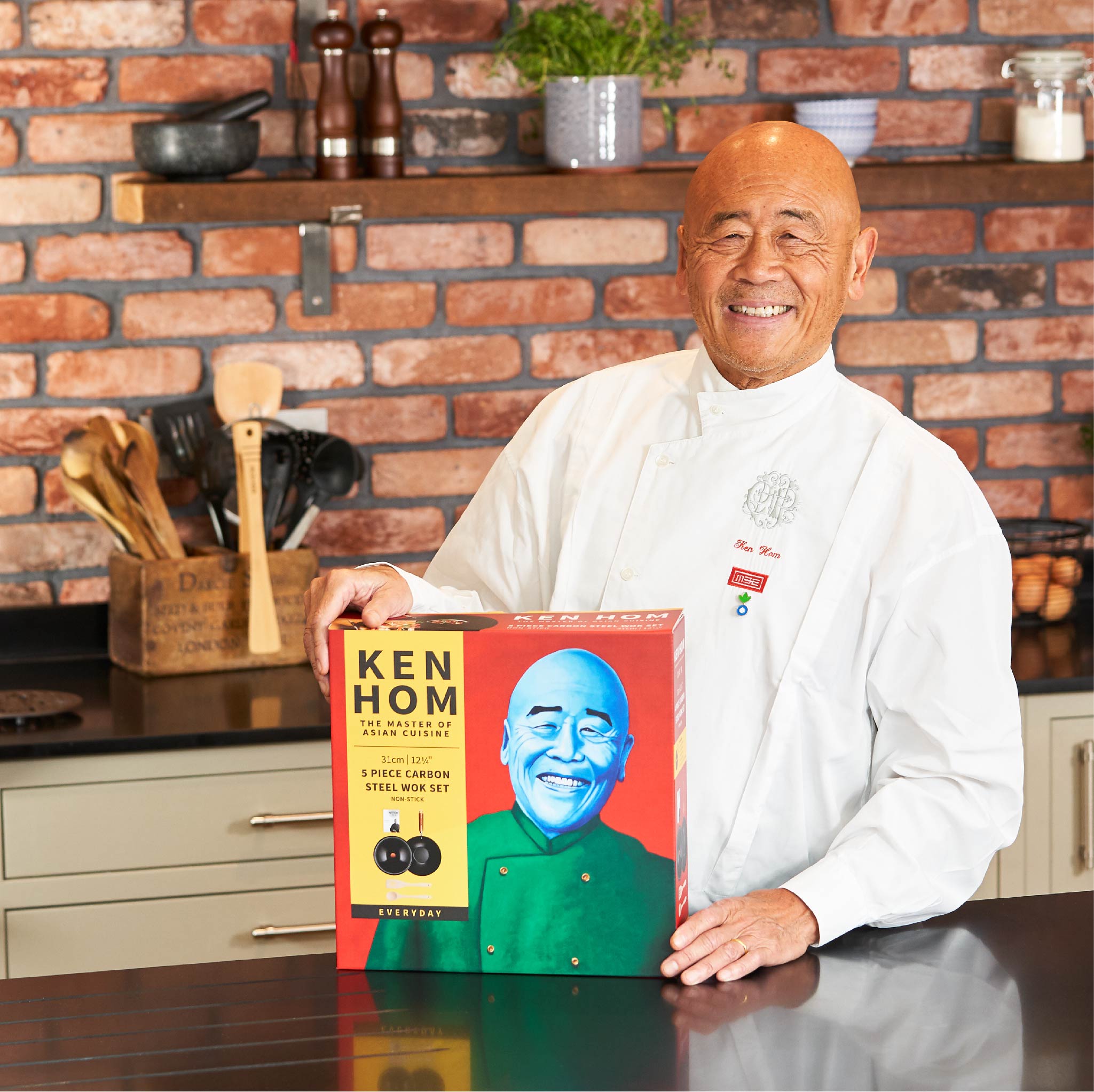 A smiling chef in a white coat stands in a kitchen, holding a boxed Ken Hom carbon steel wok set with his photo on the packaging. The kitchen has a brick wall, utensils, and plants in the background.