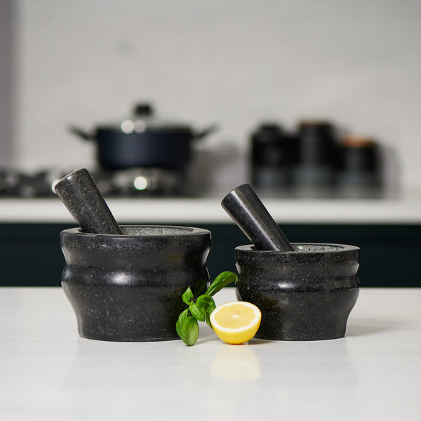 Two Cole & Mason Worcester Black Granite Pestle & Mortar sets rest on a white countertop, ideal for grinding herbs and spices. A sprig of basil and a halved lemon are in front, with a stove and kitchen items softly blurred in the background.