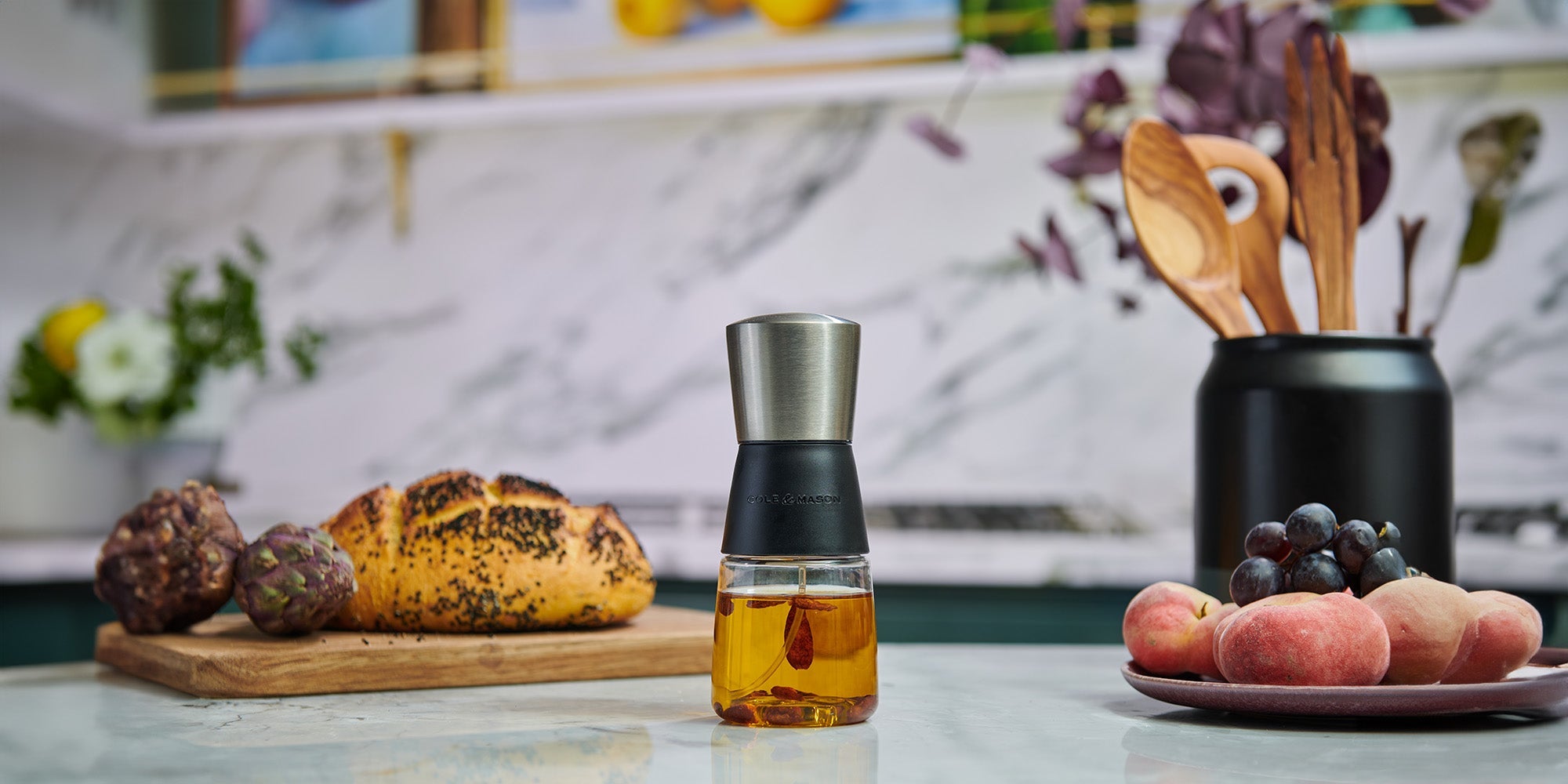 A glass container with a metal lid filled with oil sits on a kitchen counter. In the background, there is a loaf of bread, artichokes, wooden utensils in a black holder, grapes, and peaches.