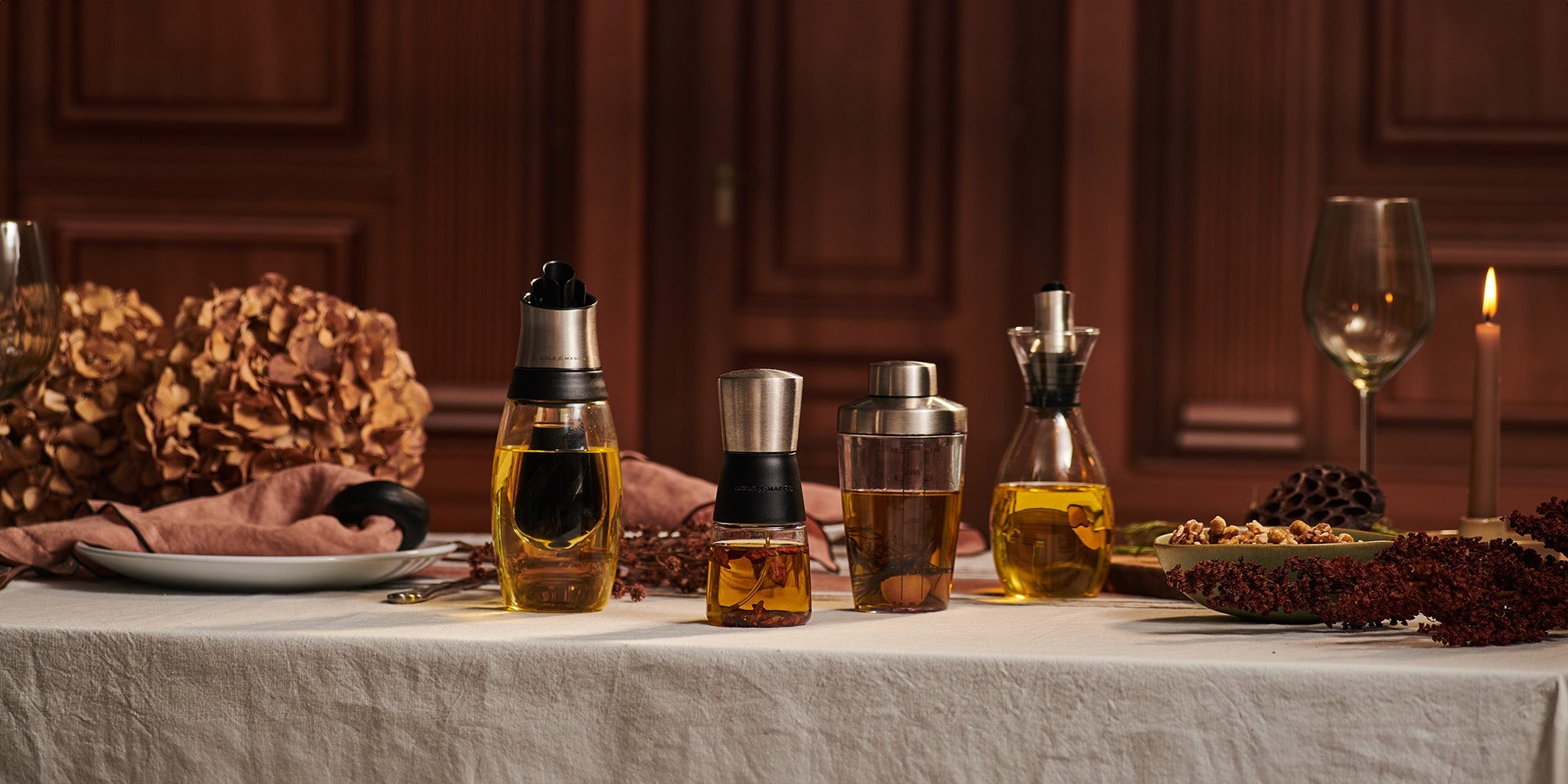 A table set with various oil and vinegar dispensers, dried flowers, a wine glass, a plate with a napkin, a burning candle, and a dish of nuts, all arranged on a white tablecloth in a warmly lit room.