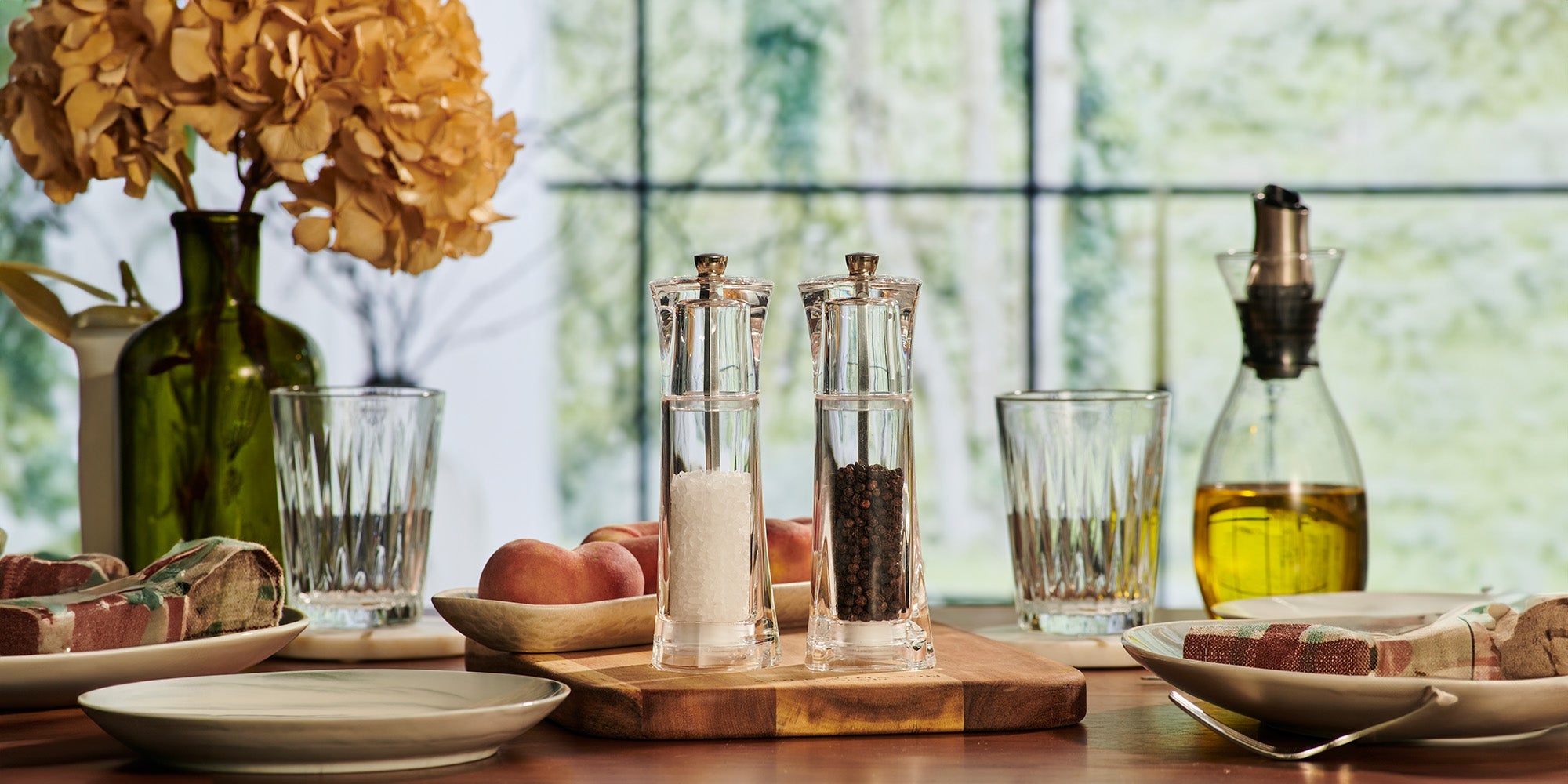 A dining table set with salt and pepper grinders, glasses, a bottle of olive oil, wrapped butter, peaches in a wooden bowl, and plates; a vase with dried flowers is in the background.
