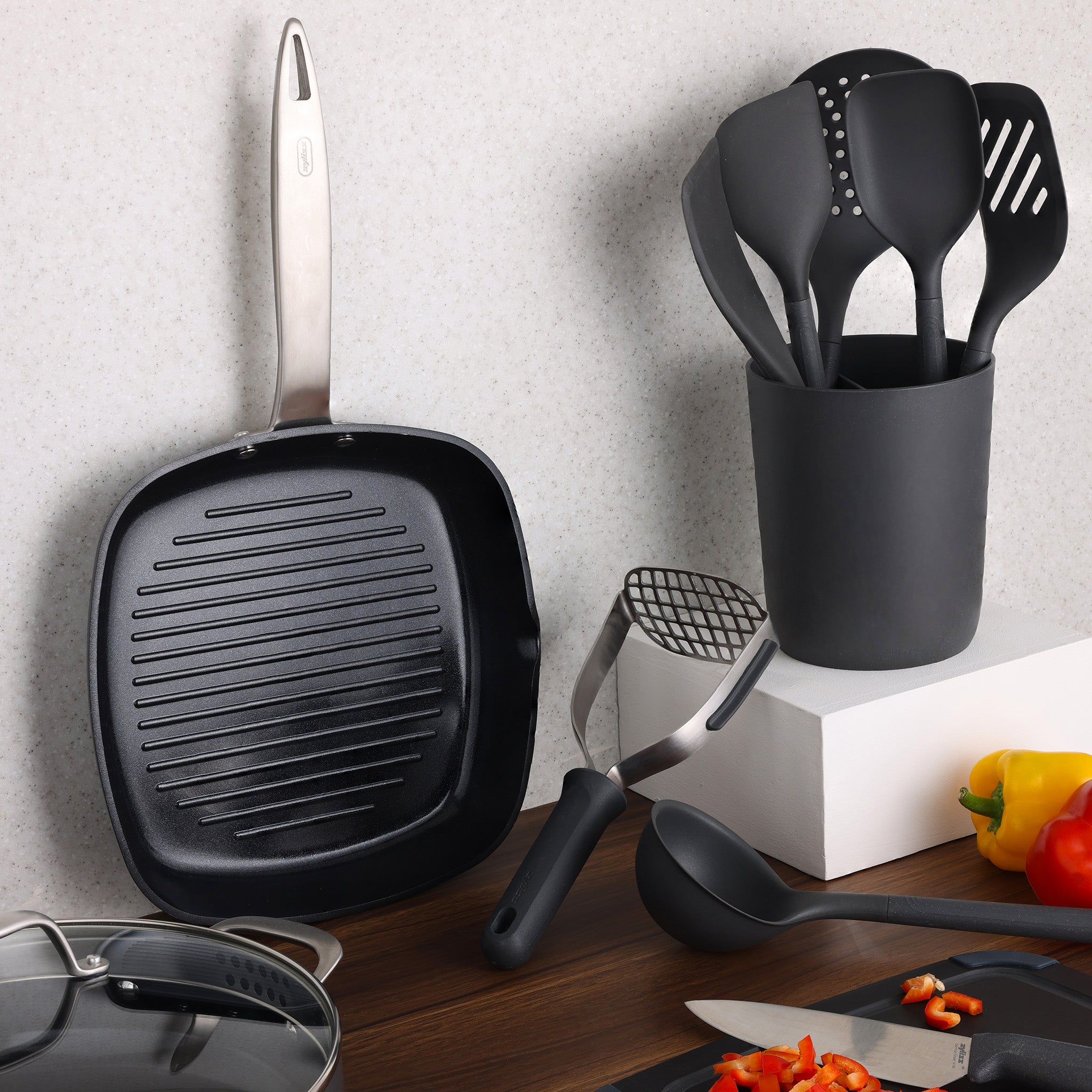 A black grill pan with a metal handle lies on its side next to a container holding black kitchen utensils. Other utensils, chopped vegetables, and a pot lid are on a wooden countertop.