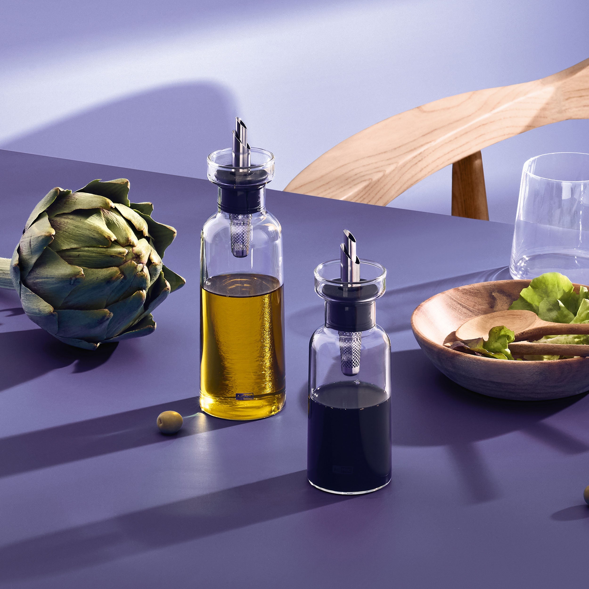 Two glass bottles with metal pour spouts filled with oil and vinegar sit on a purple table near a wooden chair, an artichoke, a glass of water, and a wooden bowl with salad greens.