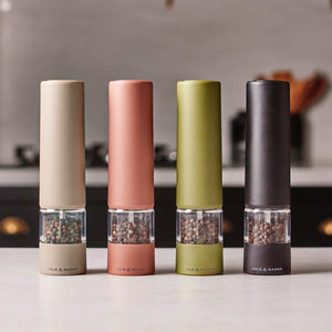 Four modern Cole & Mason Brixton Electric Mill Terracotta GS grinders—each equipped with a ceramic grinding system and available in beige, terracotta, olive green, and black—stand in a row on a kitchen countertop with a blurred background.