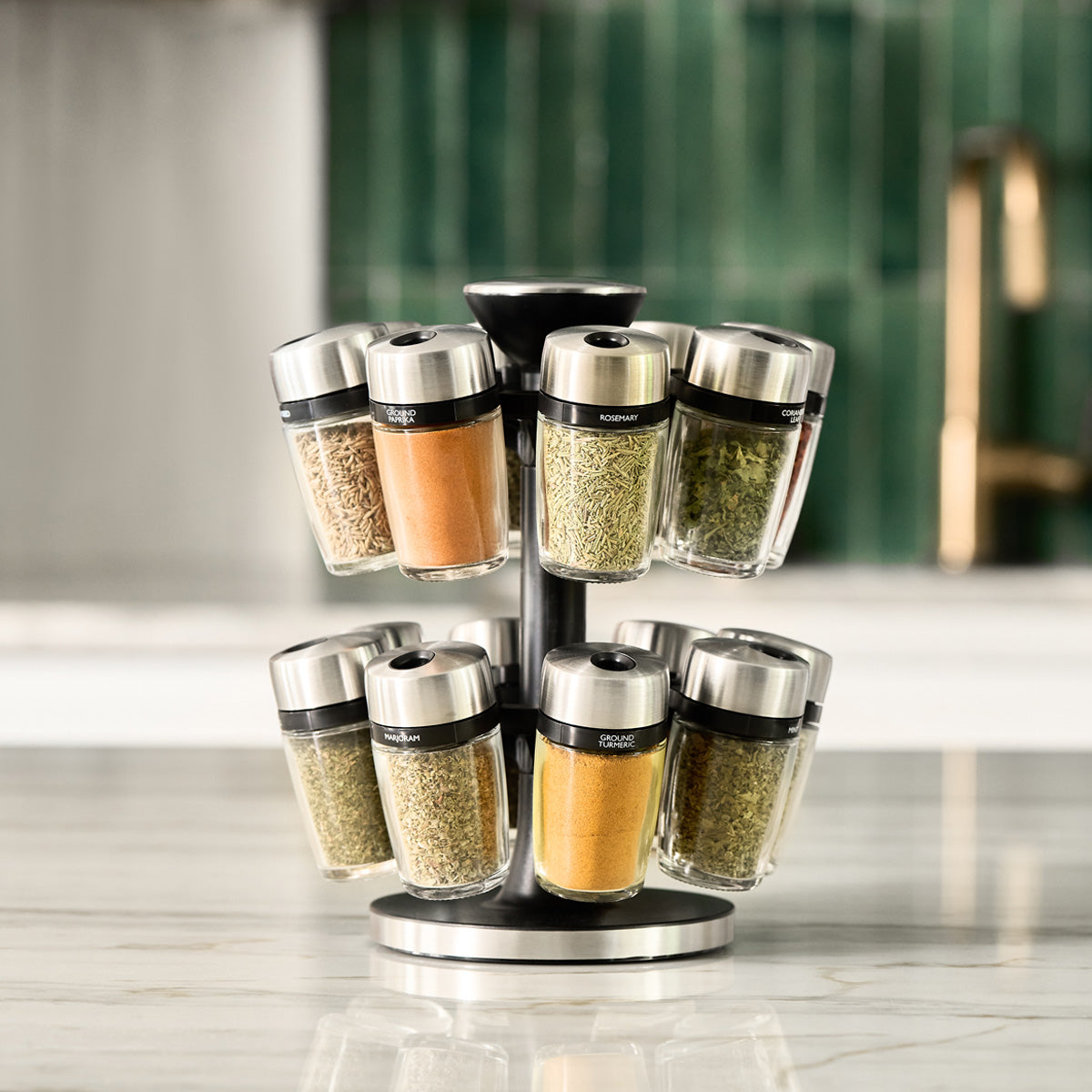A rotating spice rack with glass jars containing various spices sits on a marble countertop. The background features green tile and a blurred gold faucet.