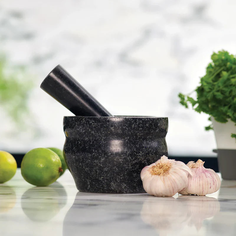A black stone mortar and pestle sit on a marble countertop with two bulbs of garlic in front, limes to the left, and a potted green herb to the right. The background is softly blurred.