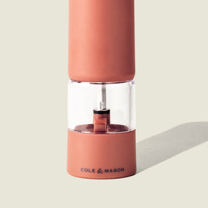 A terracotta COLE & MASON Brixton Electric Mill GS with a clear middle section displaying peppercorns inside, featuring a durable ceramic grinding system and casting a shadow on a plain light background.