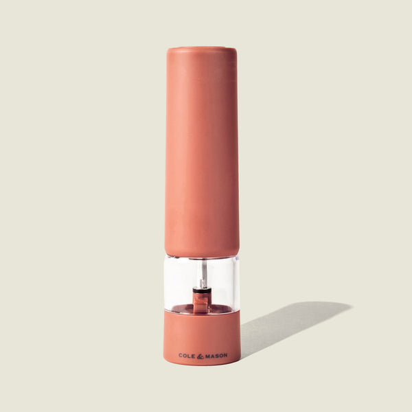 The Cole & Mason Brixton Electric Mill Terracotta GS is a tall, cylindrical rechargeable electric mill in a rose-colored finish, with a transparent middle section and Cole & Mason branding on the base. It features a ceramic grinding system and is shown against a plain light background.