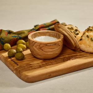 The Cole & Mason Portobello Olive Wood Salt Cellar sits open on a wooden board with coarse salt, accompanied by green olives, a colorful napkin, and seeded bread rolls for an inviting kitchen display.