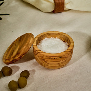 The Cole & Mason Portobello Olive Wood Salt Cellar, filled with coarse salt, sits on a beige cloth beside green olives—its lid and engraved detail make it a stylish addition to any kitchen.