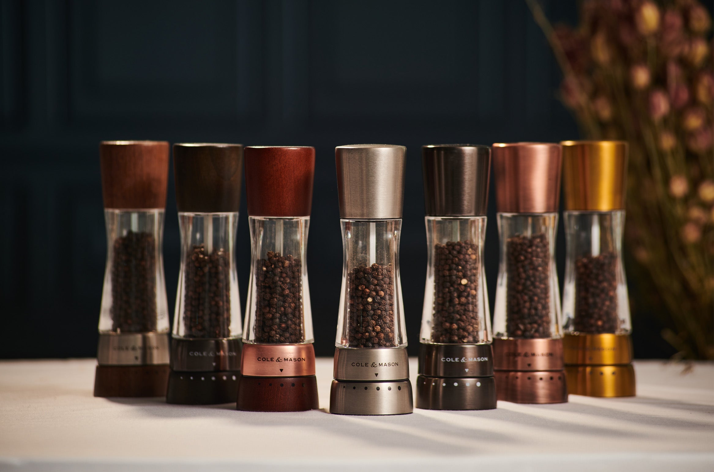 A row of six pepper grinders in various metallic and wooden finishes is displayed on a white surface, with blurred dried flowers in the background. The grinders contain whole peppercorns.