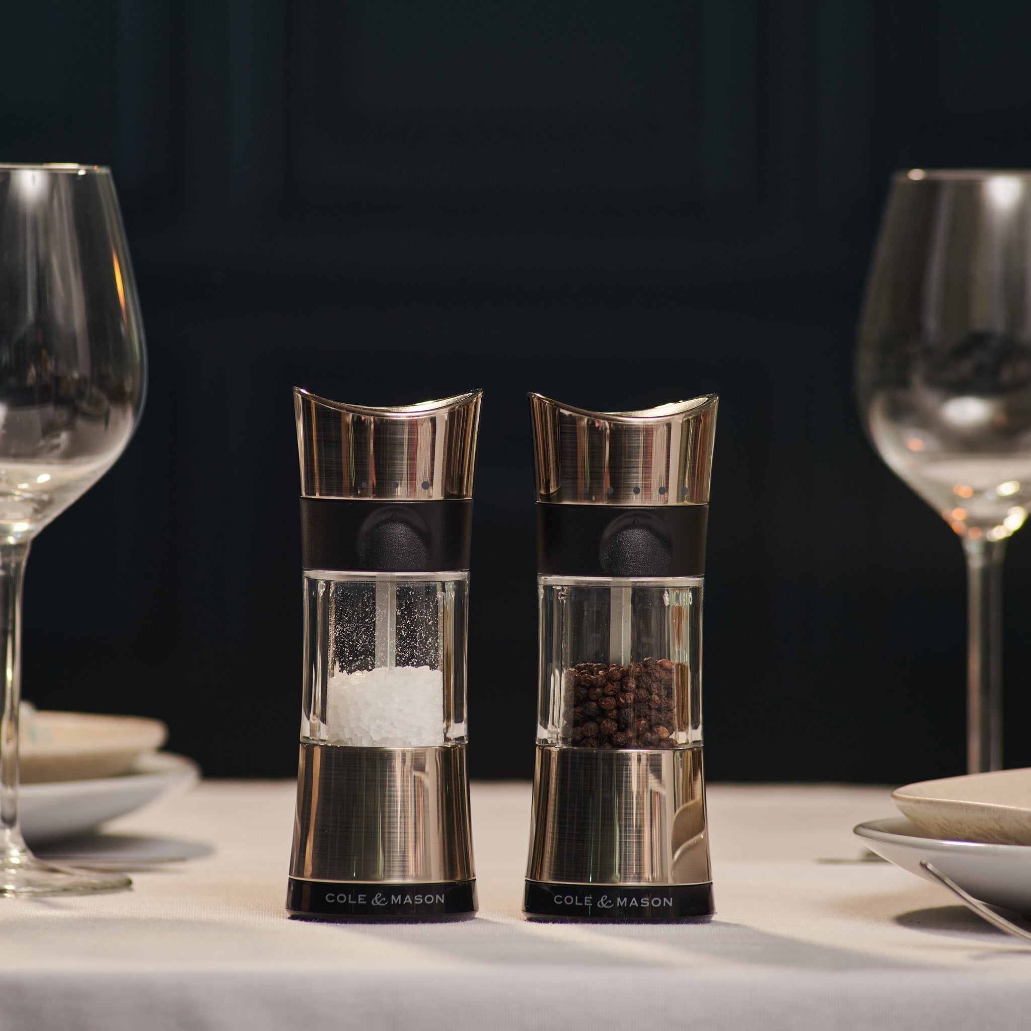 Two Cole & Mason salt and pepper grinders sit on a neatly set dining table with wine glasses, plates, and folded napkins, against a dark background.