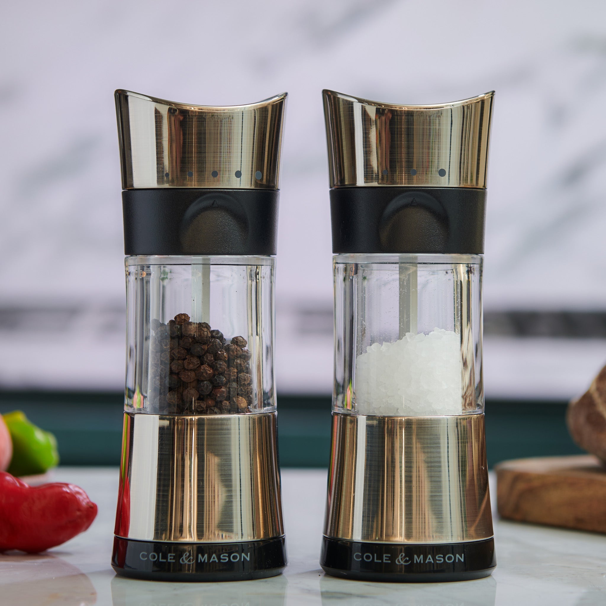 Two Cole & Mason grinders sit side by side on a kitchen counter; one is filled with black peppercorns and the other with coarse white salt. The background is out of focus, showing a pepper and a wooden board.