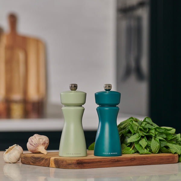 Two Cole & Mason Kenton Sage Salt & Pepper Mills, with durable ceramic mechanisms and light green and teal finishes, rest on a wooden board alongside fresh herbs and garlic, adding style to your kitchen counter.